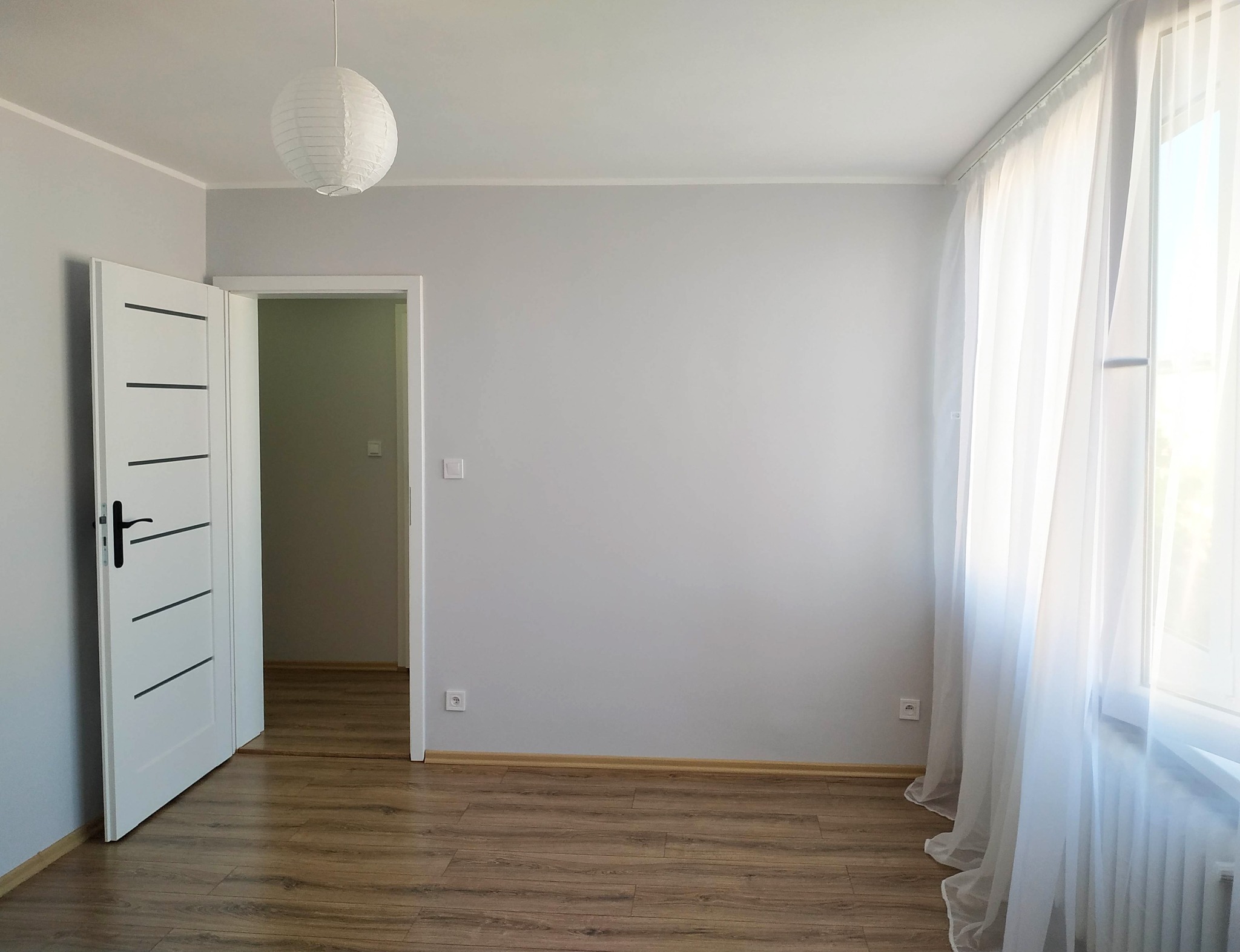 Jasne, minimalistyczne wnętrze pokoju po remoncie: świeżo pomalowane szare ściany, drewnopodobna podłoga, białe drzwi z czarnymi akcentami i zwiewne białe zasłony przy oknie.