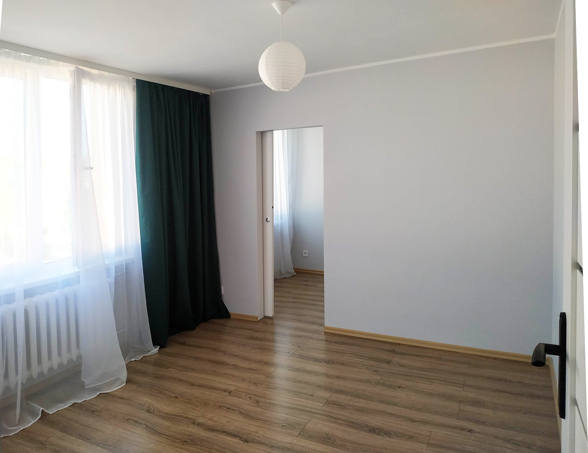 Pomalowane na szaro wnętrze pokoju z drewnianą podłogą, białymi zasłonami i ciemnozielonymi kotarami, widoczny okrągły, papierowy klosz lampy sufitowej i otwarte drzwi do sąsiedniego pomieszczenia.