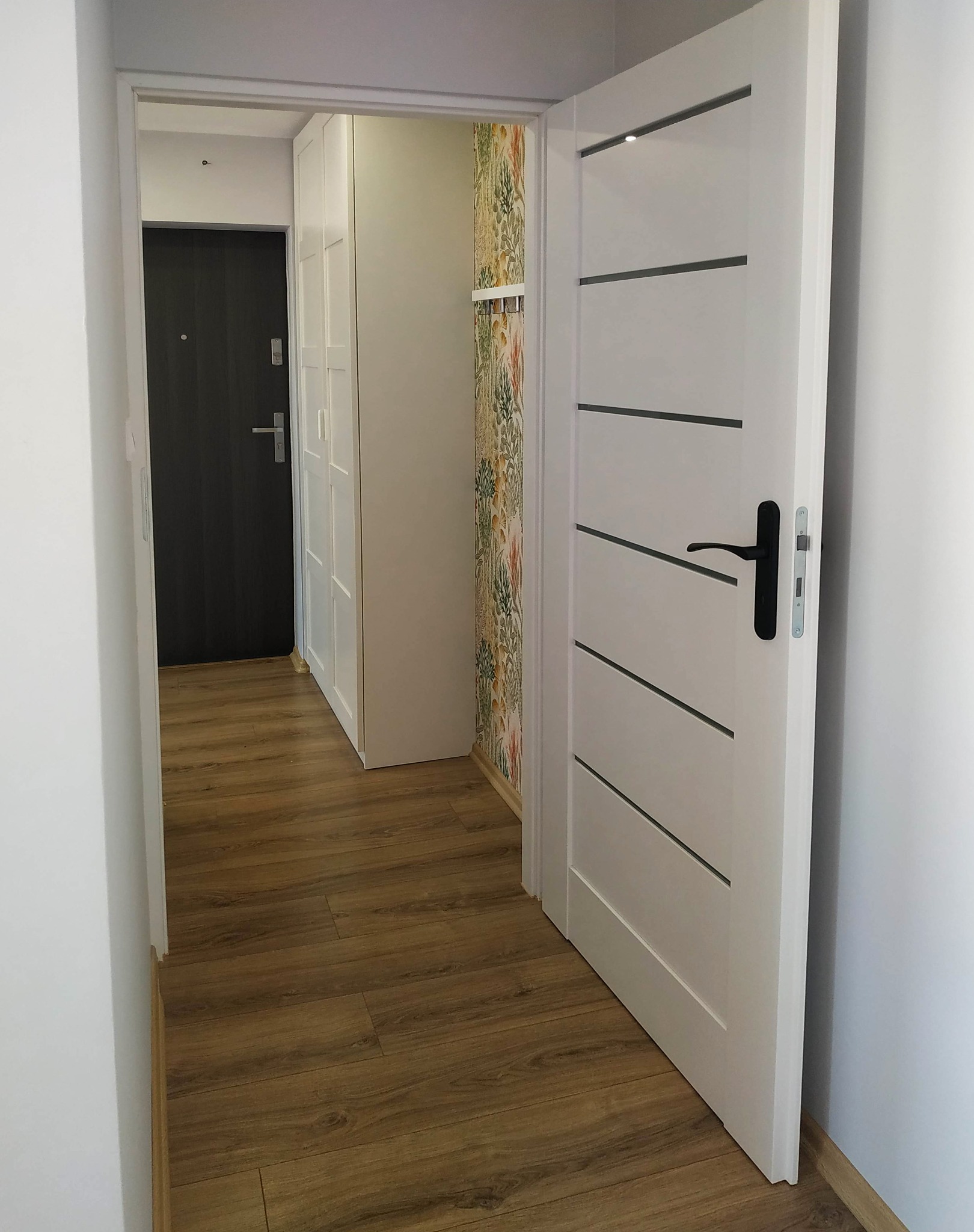 Wnętrze mieszkania po remoncie: brązowe panele podłogowe, białe drzwi wewnętrzne z czarną klamką, fragment szafy wnękowej i tapeta w roślinne wzory.