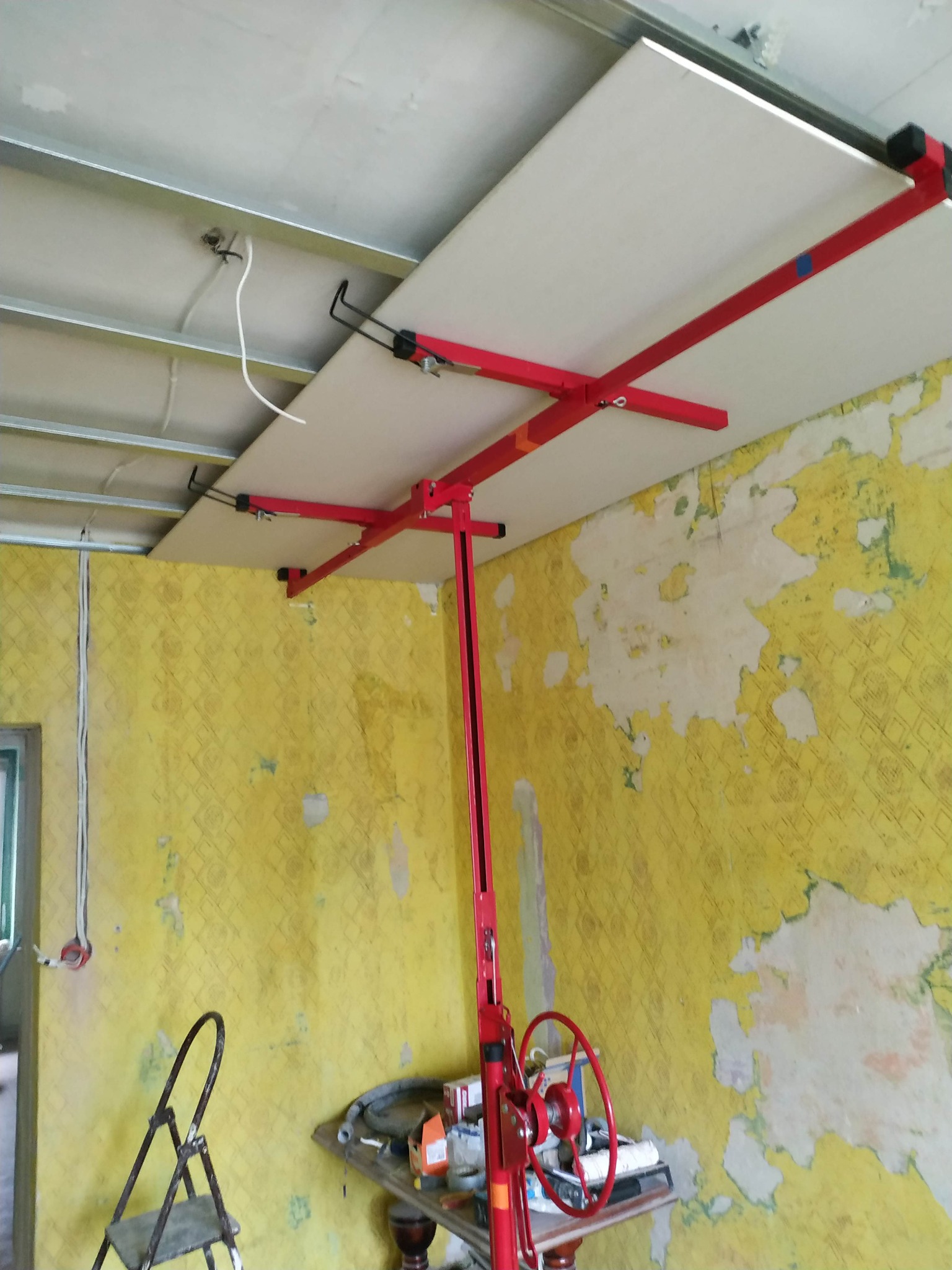 Montaż płyty gipsowo-kartonowej na suficie za pomocą podnośnika, widoczne metalowe profile sufitowe i fragmenty starej, żółtej tapety na ścianach.
