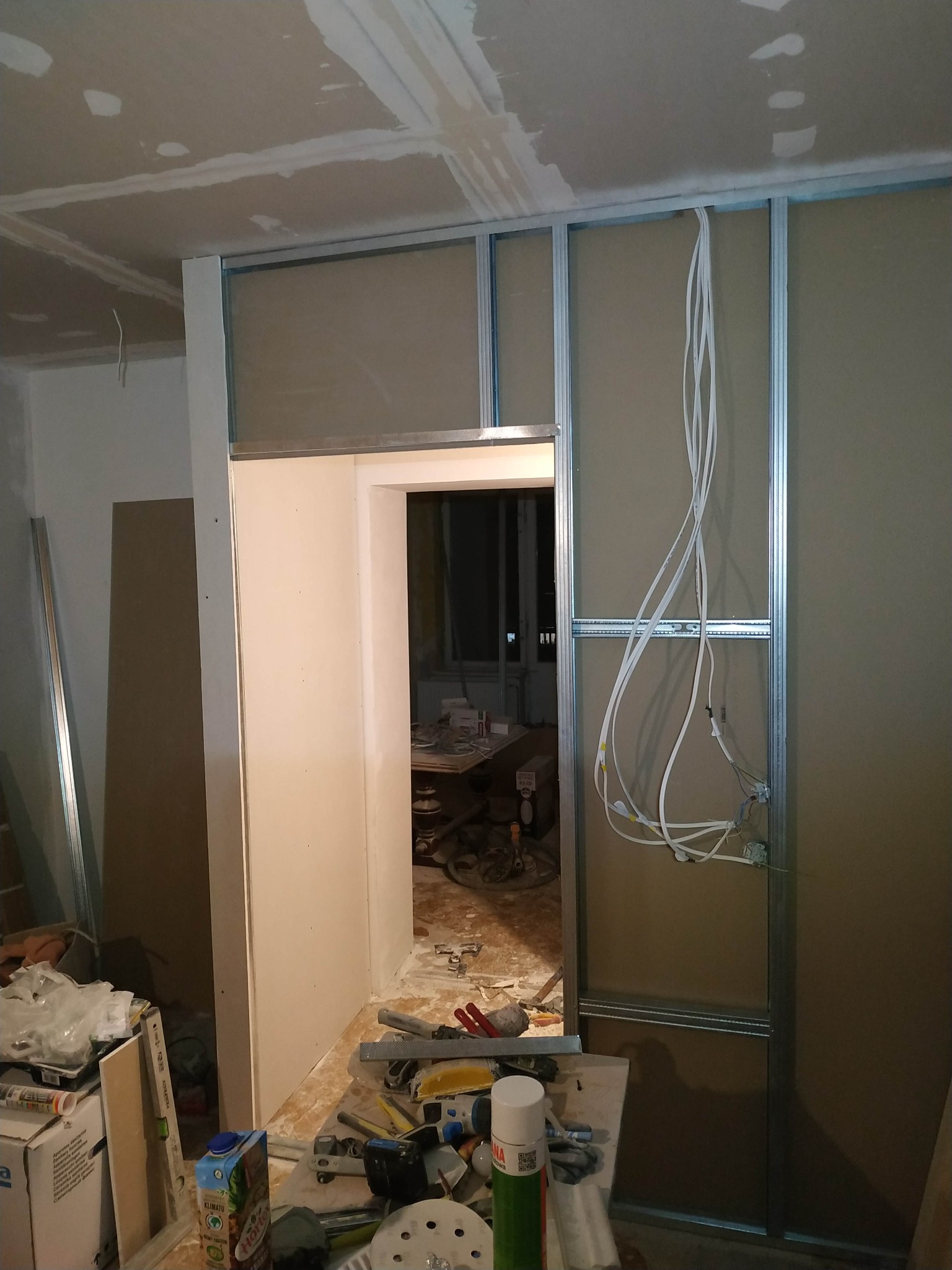 Wnętrze w trakcie prac: montaż stelaża z płyt gipsowo-kartonowych, widoczne przewody elektryczne, narzędzia i materiały budowlane na podłodze.