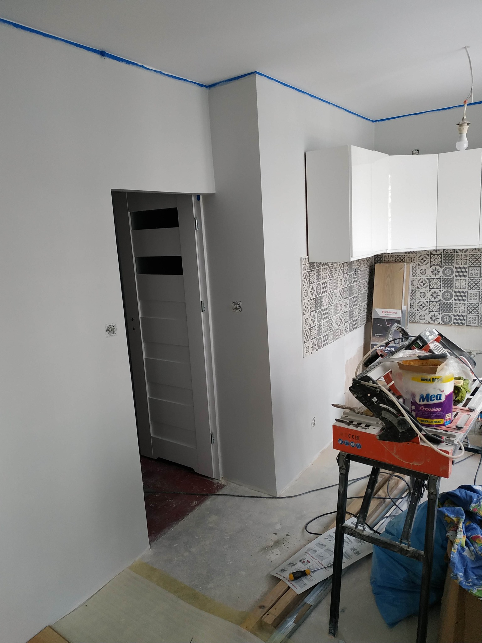 Wnętrze mieszkania w trakcie prac remontowych: świeżo pomalowane białe ściany zabezpieczone niebieską taśmą malarską przy suficie, widoczna kuchnia z białymi szafkami i wzorzystą glazurą,...