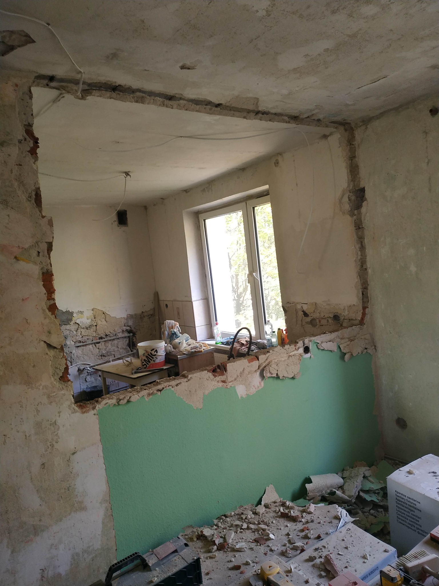 Wnętrze w trakcie prac remontowych: wyburzona ściana, odsłonięte cegły, fragment zielonej ściany, gruz, narzędzia i wiadro farby widoczne w tle, okno z widokiem na zieleń.