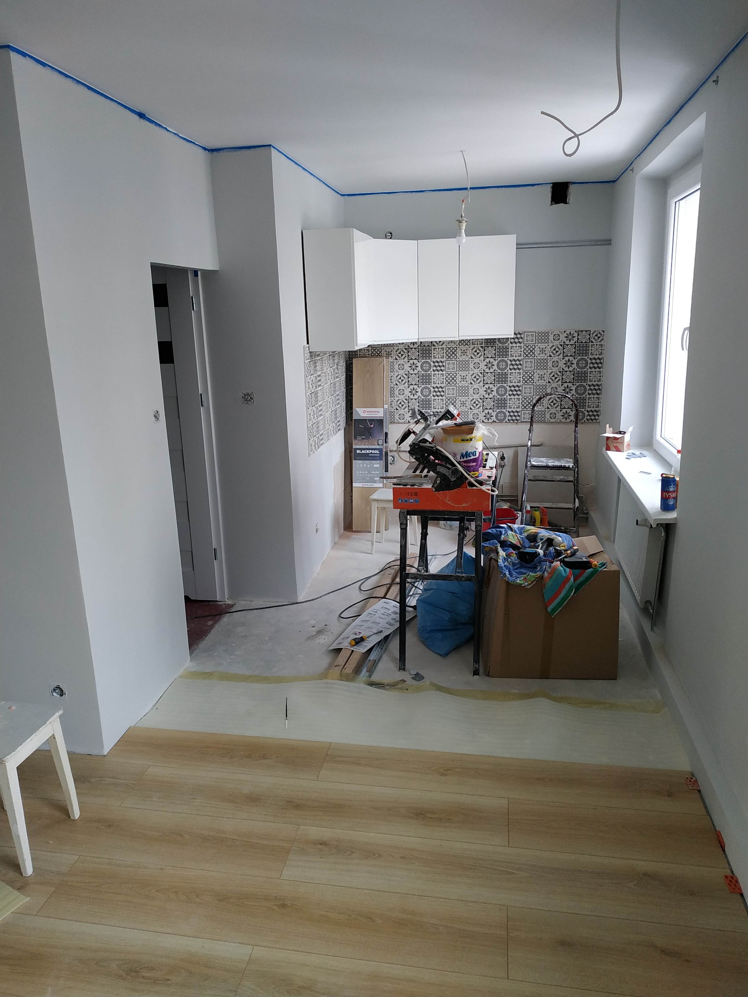 Wnętrze w trakcie prac wykończeniowych: ułożona podłoga z paneli, ściany przygotowane do malowania z niebieską taśmą, widoczna kuchnia z mozaiką na ścianie i narzędzia glazurnicze na stole.