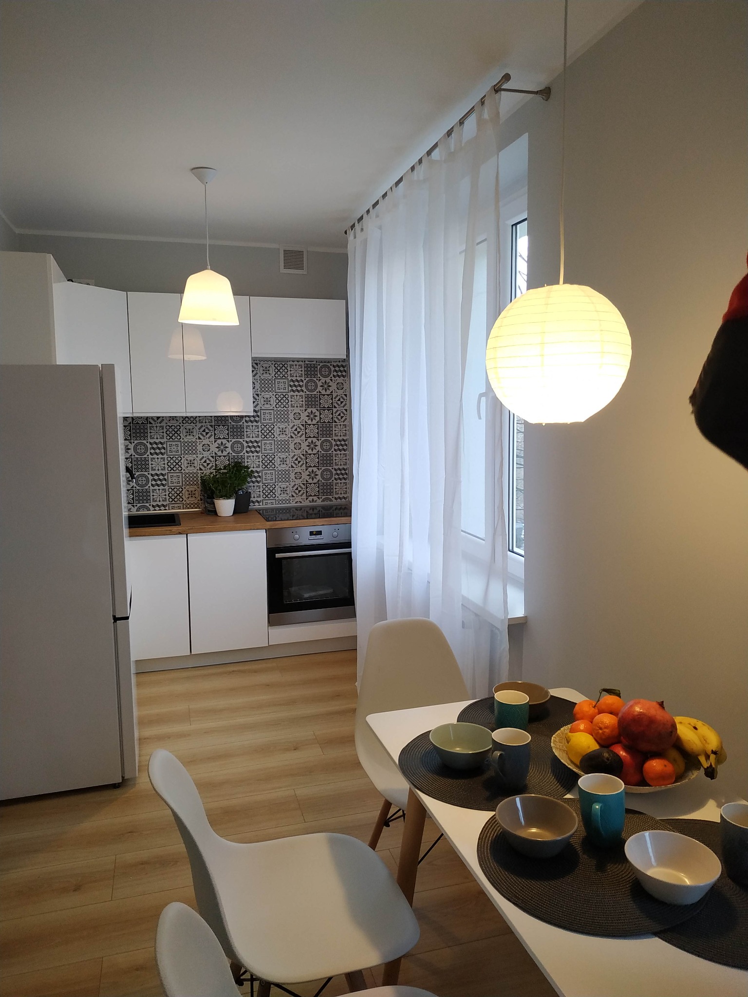 Jasne wnętrze kuchni z jadalnią, białe meble, drewniany blat, mozaika nad blatem, stół z owocami i nakryciem, lampy wiszące.