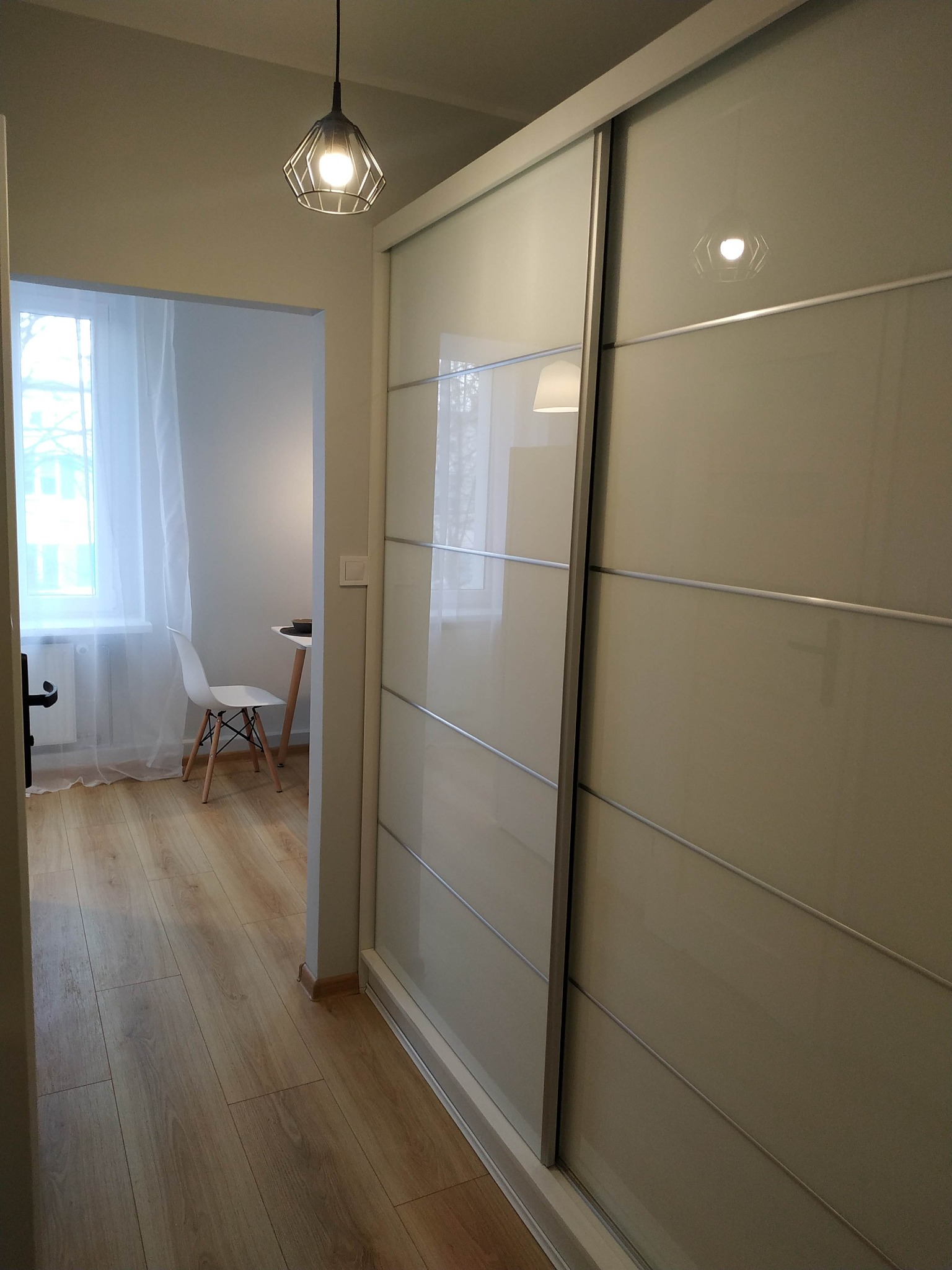 Jasne panele podłogowe w korytarzu prowadzącym do pokoju z białym krzesłem i stołem, obok duża szafa przesuwna z jasnymi frontami.