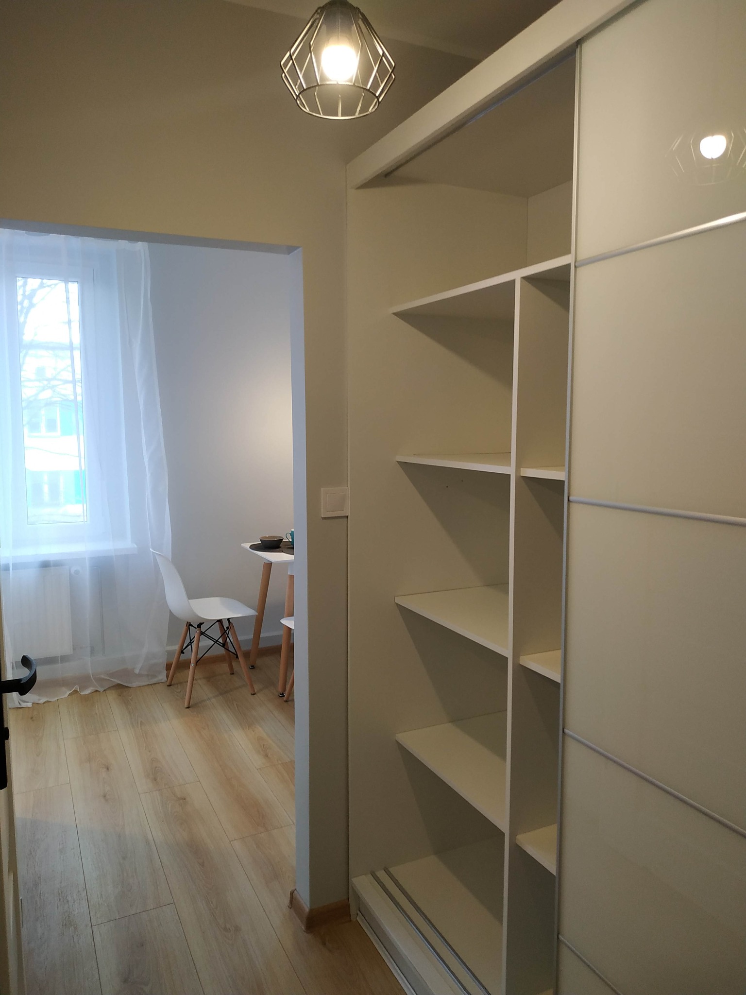 Jasne wnętrze z widokiem na pokój z oknem i białym stołem, w tle biała szafa przesuwna z półkami.