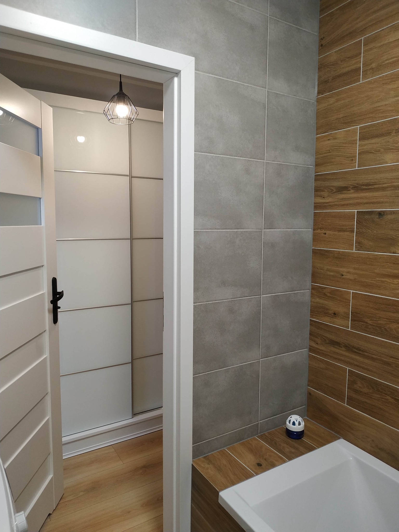 Wykończona łazienka z szarymi i drewnopodobnymi płytkami, białą wanną i otwartymi drzwiami do garderoby z przesuwnymi drzwiami, widoczna lampa sufitowa w stylu industrialnym.