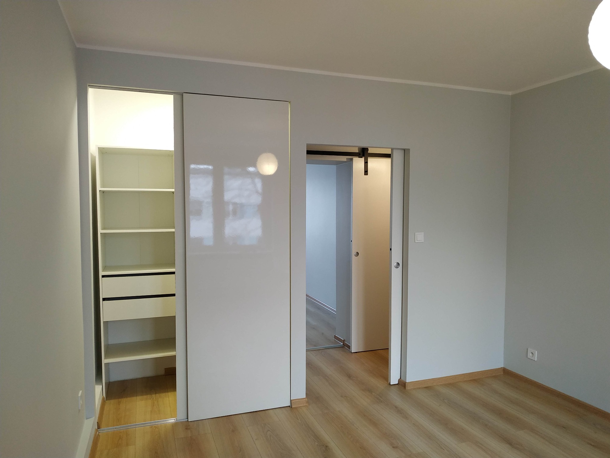 Wnętrze pokoju po remoncie z jasnymi ścianami, podłogą z paneli i dwiema parami przesuwnych drzwi: jedne do garderoby z półkami i szufladami, drugie do innego pomieszczenia z widoczną szyną.