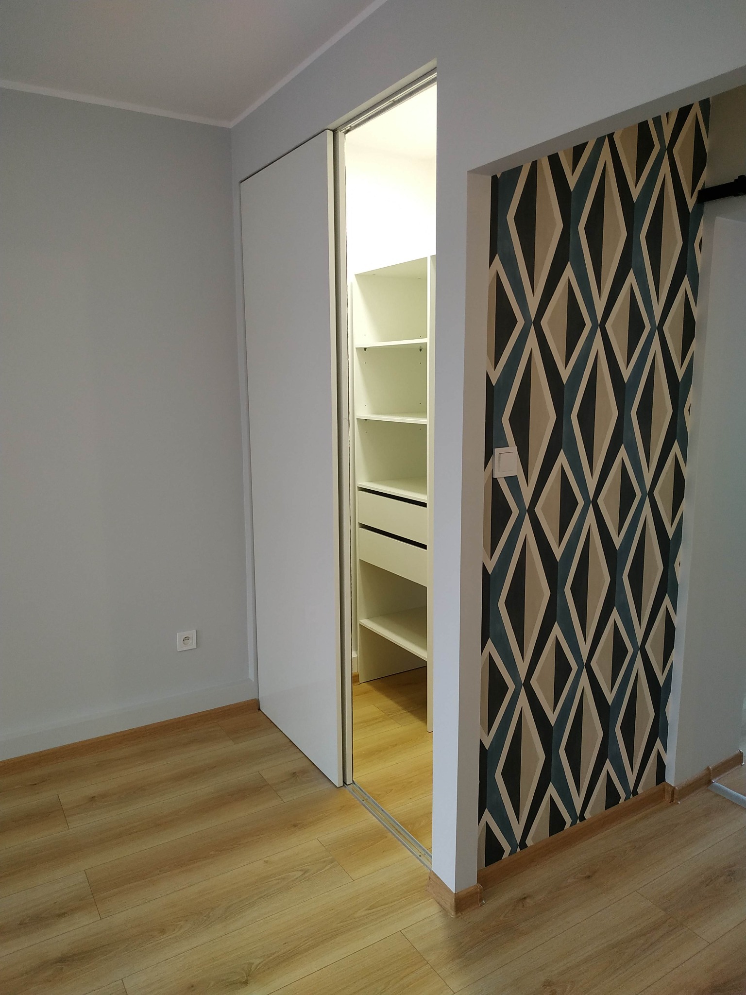 Wnętrze pokoju z drewnianą podłogą, szarą ścianą i białą szafą wnękową z przesuwnymi drzwiami, obok ściana z geometryczną tapetą w odcieniach szarości, beżu i błękitu.
