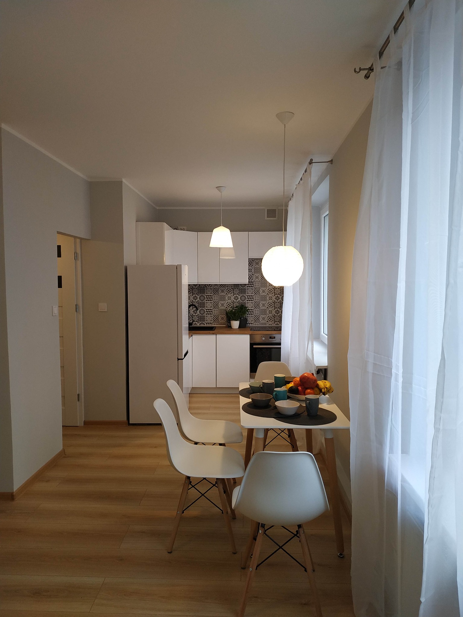 Jasne mieszkanie z drewnianą podłogą, białymi ścianami i kuchnią w tle, stół z owocami i naczyniami w jadalni, widoczne białe zasłony przy oknie.