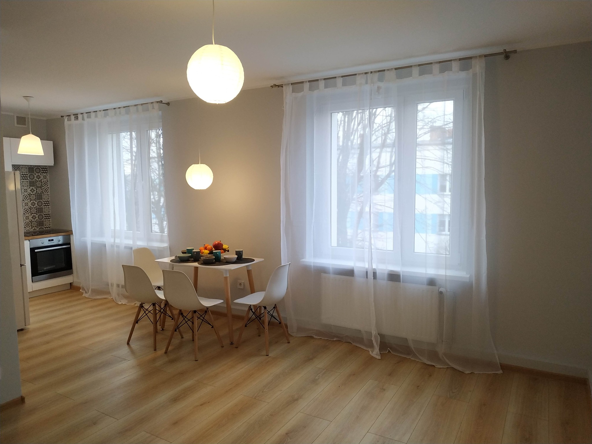 Jasne, wykończone wnętrze mieszkania w Laryszowie z parkietem, białymi ścianami, oknami z białymi firankami i nowoczesnym oświetleniem, ukazujące stół z owocami i krzesłami.