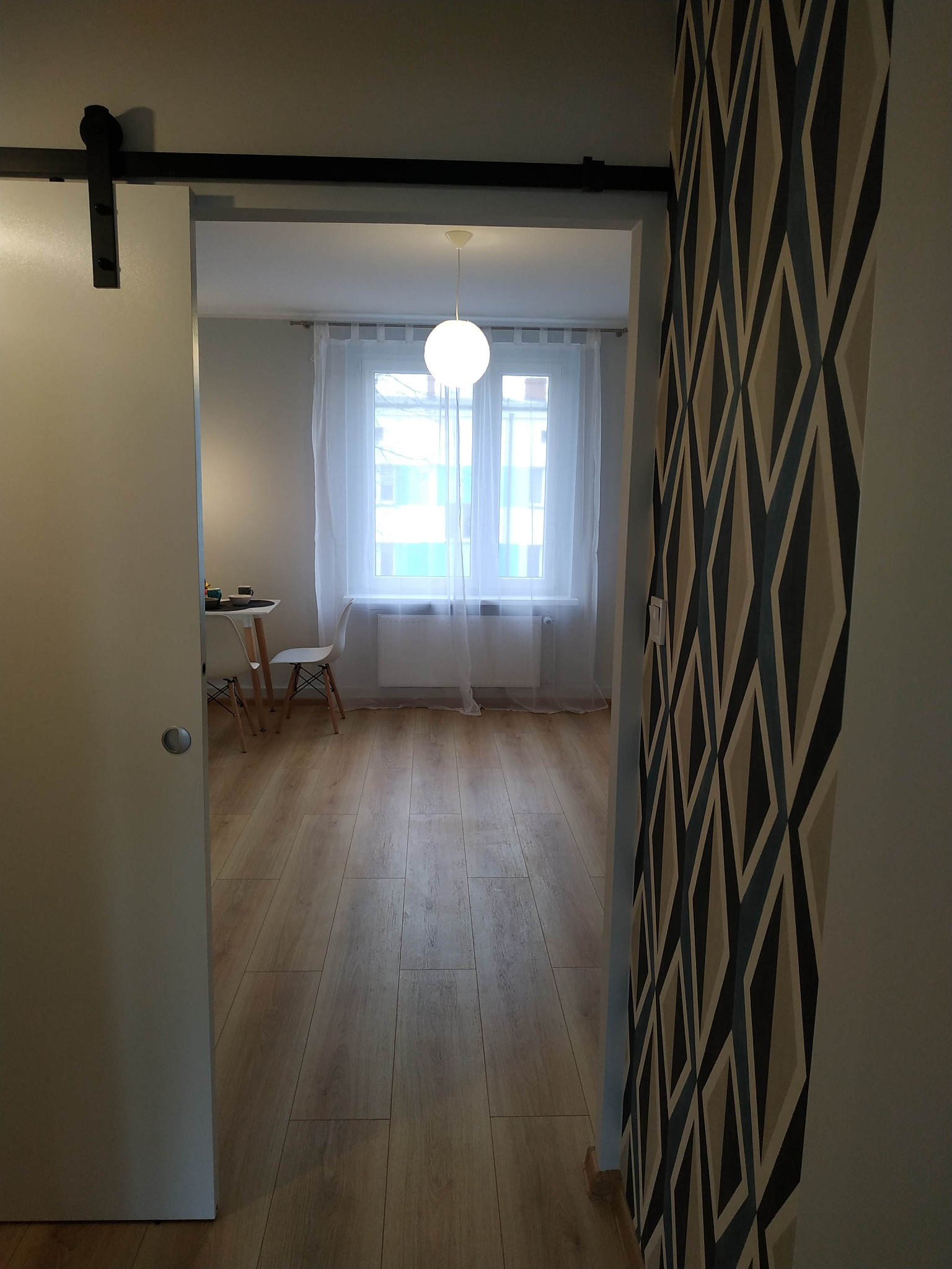 Wnętrze mieszkania z drewnianą podłogą, widok przez przesuwne drzwi z czarnym systemem szynowym, akcentująca ściana z geometrycznym wzorem tapety.