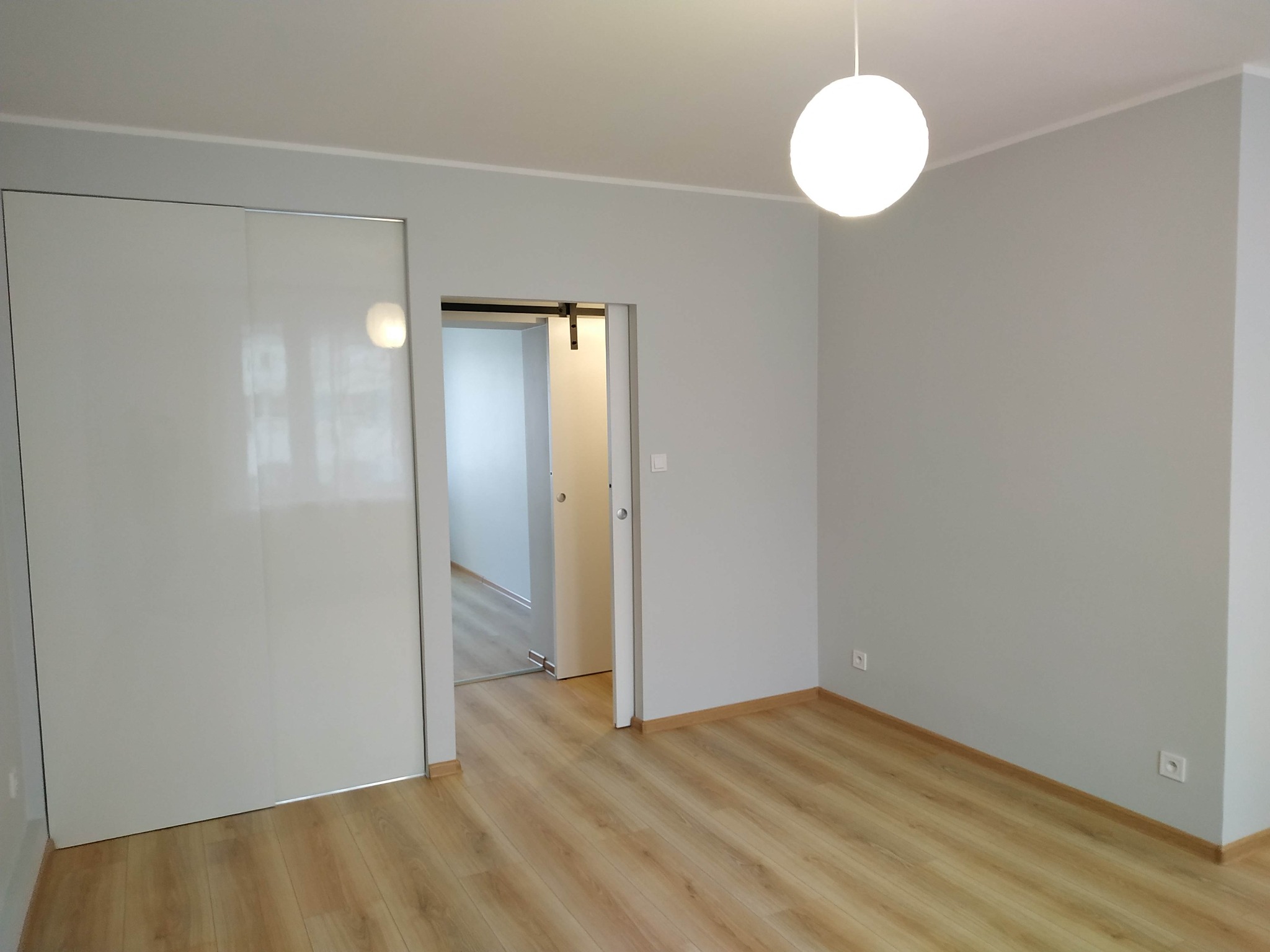 Pusty pokój z jasnymi panelami podłogowymi, szarą ścianą, przesuwnymi białymi drzwiami szafy i otwartymi drzwiami do sąsiedniego pomieszczenia, oświetlony kulistą lampą wiszącą.