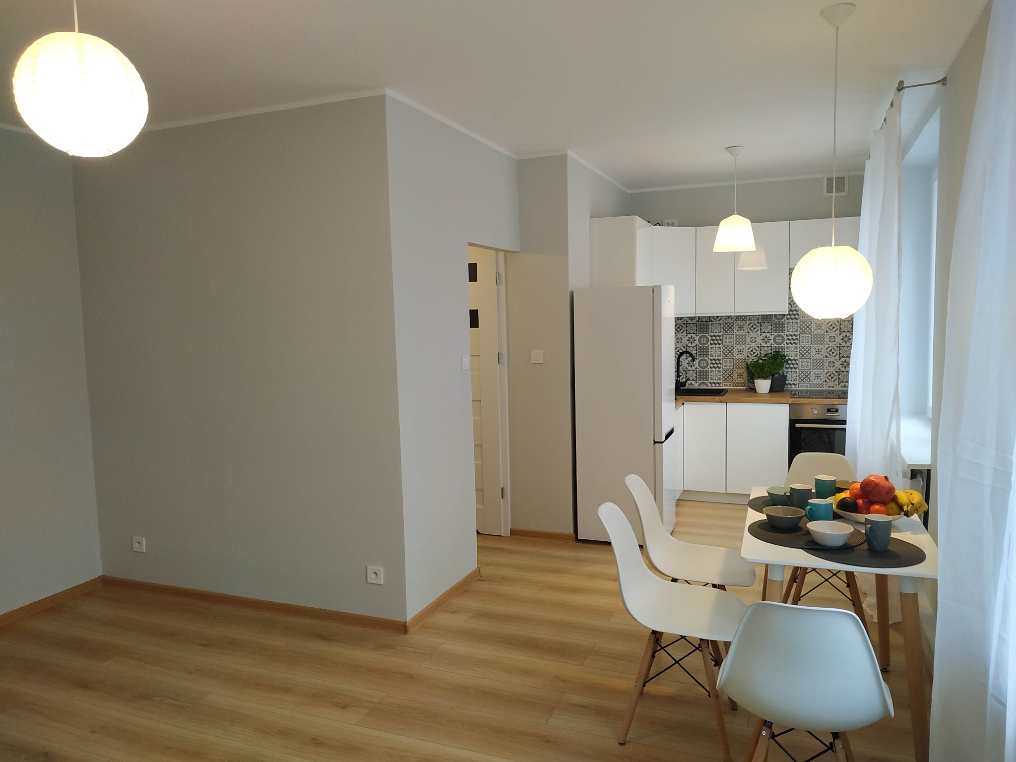 Jasne wnętrze mieszkania po remoncie: szare ściany, drewniana podłoga, białe meble kuchenne i stół z owocami, oświetlone wiszącymi lampami.