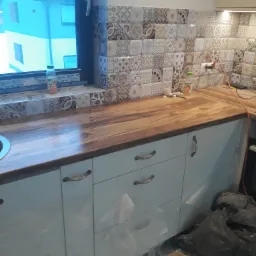 Nowa drewniana płyta blatu kuchennego z mozaiką płytek na ścianie, widok z boku na białe szafki z metalowymi uchwytami, butelka soku na parapecie okna.