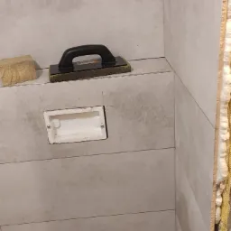 Łazienka w trakcie wykańczania: szare, poziome płytki, narzędzie glazurnicze, puszka instalacyjna, rury. Widoczne detale wykończeniowe i etap pracy.