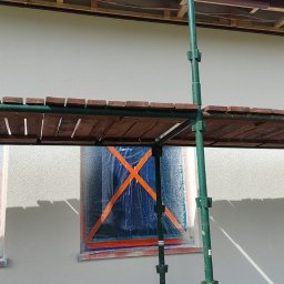 Usługi Wykończenia Wnętrz Remontowe Ogólnobudowlane Stolarskie Ciesielskie Handyman Marek Gąsiewicz - Elewacja domu w trakcie malowania, okna zabezpieczone folią z pomarańczową taśmą, widoczne rusztowanie. Jasna elewacja, częściowo ocieniona.