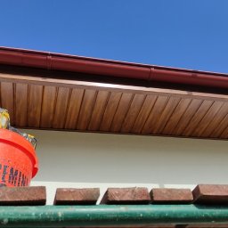 Usługi Wykończenia Wnętrz Remontowe Ogólnobudowlane Stolarskie Ciesielskie Handyman Marek Gąsiewicz - Podbitka dachowa z brązowym orynnowaniem, widok z rusztowania z wiadrem i narzędziami. Błękitne niebo w tle. Detal wykończenia elewacji.