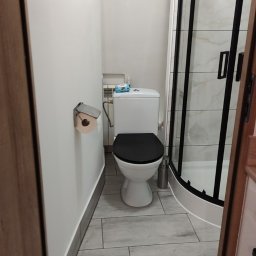 Usługi Wykończenia Wnętrz Remontowe Ogólnobudowlane Stolarskie Ciesielskie Handyman Marek Gąsiewicz - Nowoczesna łazienka z kabiną prysznicową typu walk-in, toaletą z czarną deską i szarą podłogą imitującą drewno. Widok zza drzwi.
