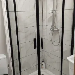 Narożna kabina prysznicowa z czarnymi profilami i marmurowymi płytkami w tle. Nowoczesne wykończenie łazienki po remoncie z widoczną armaturą.