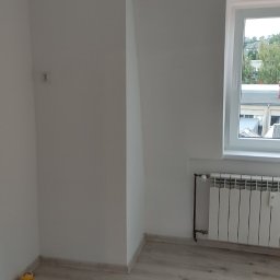 Usługi Wykończenia Wnętrz Remontowe Ogólnobudowlane Stolarskie Ciesielskie Handyman Marek Gąsiewicz - Jasne wnętrze po remoncie: białe ściany z grzejnikiem pod oknem, panele podłogowe. Widoczny mały, żółty pojemnik w rogu pokoju i elementy instalacji elektrycznej.