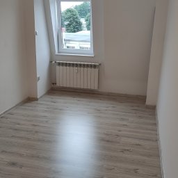 Usługi Wykończenia Wnętrz Remontowe Ogólnobudowlane Stolarskie Ciesielskie Handyman Marek Gąsiewicz - Jasne wnętrze po remoncie: panele podłogowe, białe ściany, okno dachowe z widokiem na zieleń. Nowy grzejnik pod oknem. Minimalistyczny, odświeżony pokój.