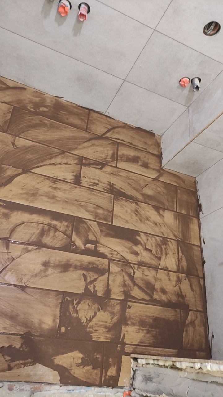 Ściana w trakcie kładzenia brązowych płytek imitujących drewno, widoczne ślady zaprawy, szare płytki na suficie i niezakończone prace wykończeniowe.