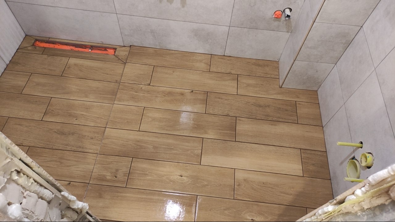 Ukończona podłoga w łazience wyłożona brązowymi płytkami imitującymi drewno, z widocznym odpływem liniowym i fragmentem ściany z szarych płytek.