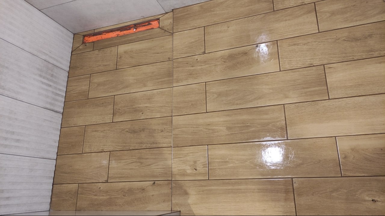 Ściana wyłożona błyszczącymi, podłużnymi płytkami imitującymi drewno w odcieniach brązu i beżu, z widocznym odpływem liniowym pod sufitem.