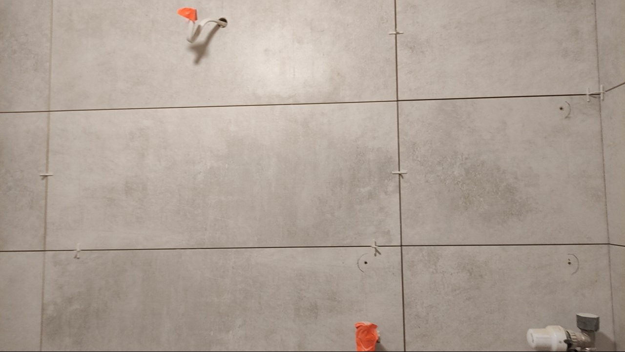 Ściana wyłożona szarymi, matowymi płytkami z krzyżykami dystansowymi i widocznymi przyłączami wodno-kanalizacyjnymi, w trakcie prac wykończeniowych.