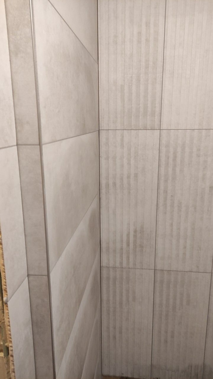 Wykończony narożnik łazienki z szarymi, prostokątnymi płytkami o zróżnicowanej fakturze. Pionowe żłobienia na jednej ze ścian. Metalowa listwa wykończeniowa.