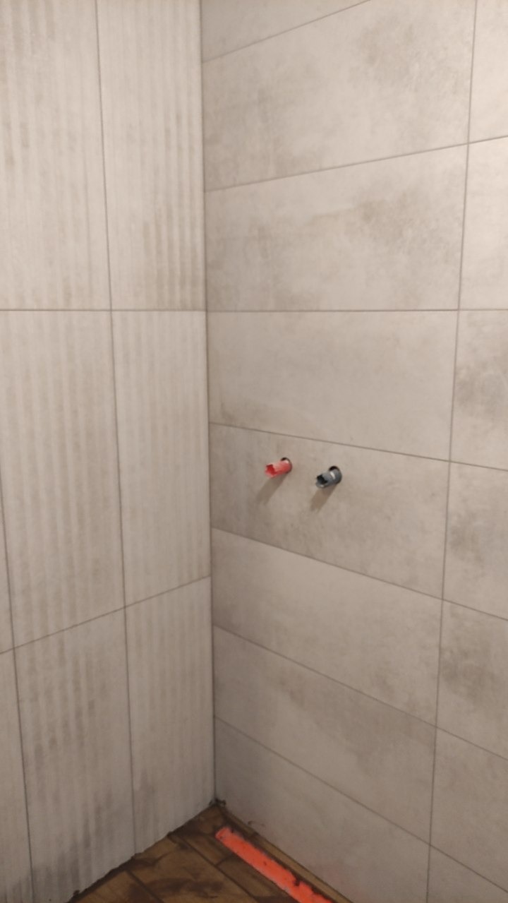 Wykończony fragment łazienki z beżowymi płytkami i wystającymi rurami. Widoczne pionowe i poziome ułożenie płytek. Podłoga wymaga wykończenia.