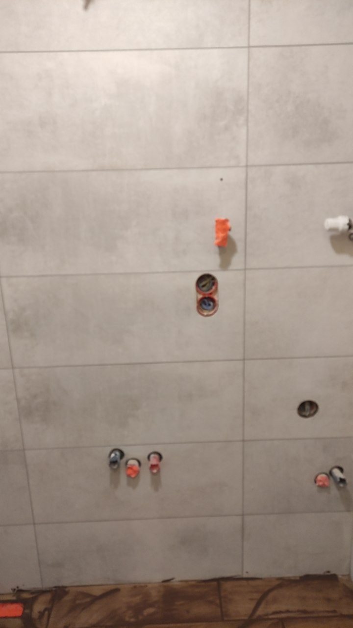 Ściana łazienki z szarymi płytkami, widoczne otwory na instalacje wodne i elektryczne. Etap prac wykończeniowych, przygotowanie do montażu armatury.