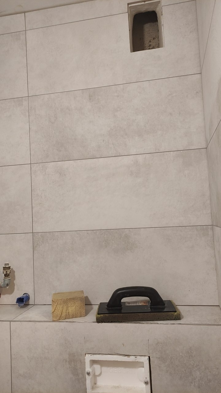 Ściana w łazience wyłożona jasnymi, poziomymi płytkami. Widoczne dwa otwory w ścianie, gąbka i packa do fugowania. Fragment instalacji wodnej po lewej.
