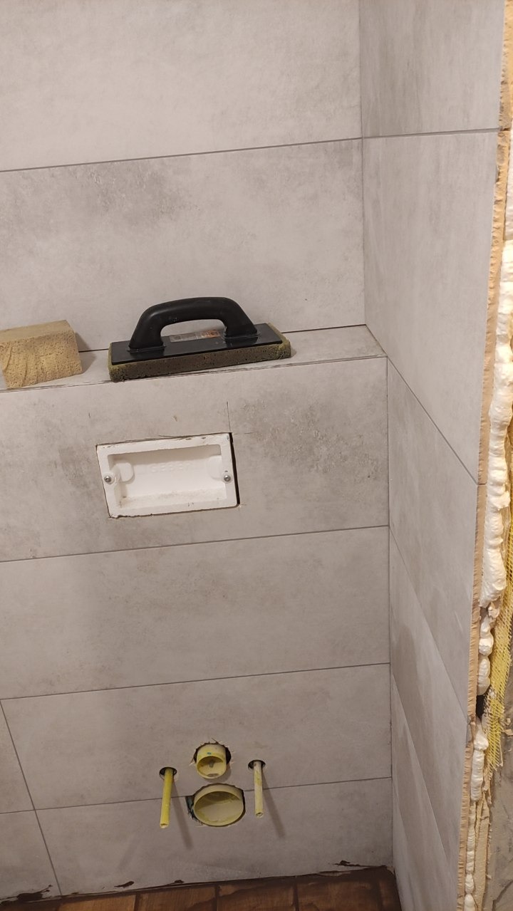 Łazienka w trakcie wykańczania: szare, poziome płytki, narzędzie glazurnicze, puszka instalacyjna, rury. Widoczne detale wykończeniowe i etap pracy.