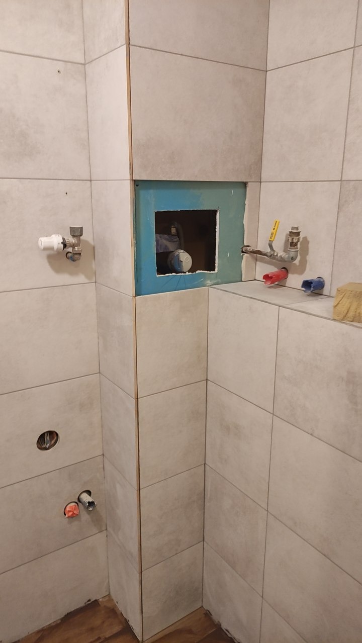 Łazienka w trakcie wykańczania: ułożone beżowe płytki ścienne, otwór rewizyjny z instalacją, widoczne rury i przyłącza. Etap prac wykończeniowych.