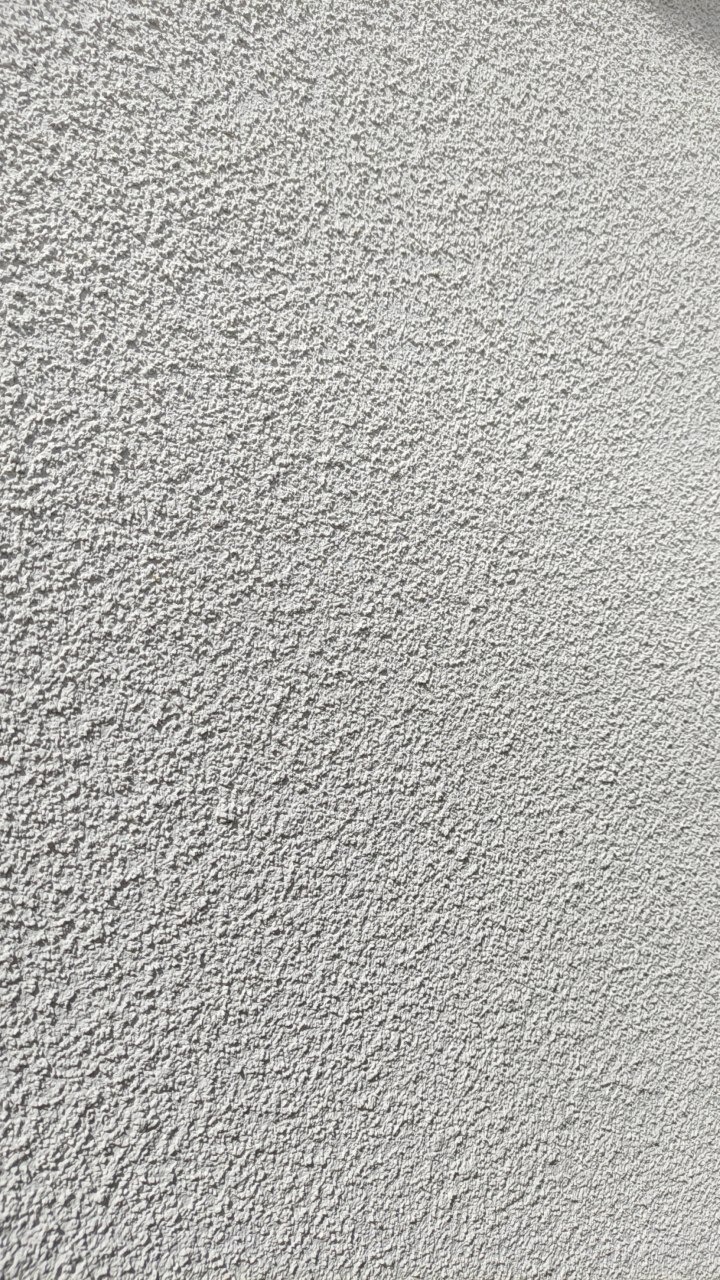 Szara, chropowata faktura ściany zewnętrznej w zbliżeniu, oświetlona z góry, ukazująca detale tynku strukturalnego w neutralnej tonacji.