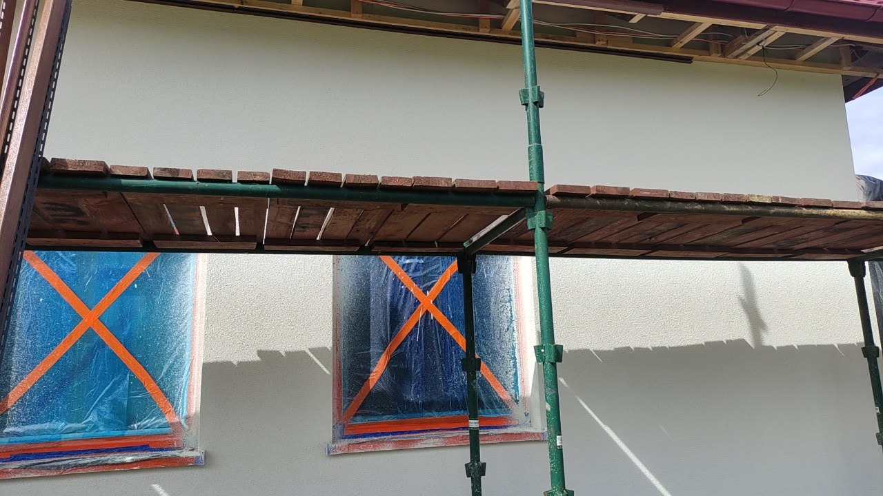 Elewacja domu w trakcie malowania, okna zabezpieczone folią z pomarańczową taśmą, widoczne rusztowanie. Jasna elewacja, częściowo ocieniona.