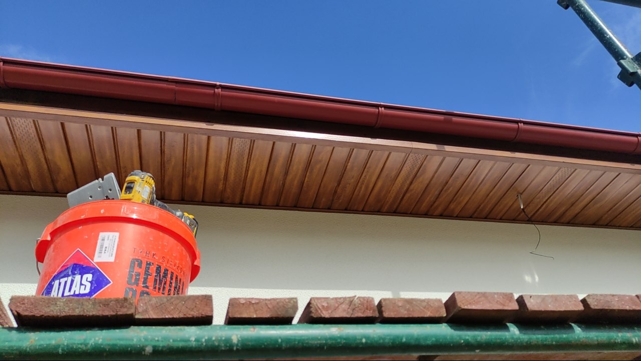 Podbitka dachowa z brązowym orynnowaniem, widok z rusztowania z wiadrem i narzędziami. Błękitne niebo w tle. Detal wykończenia elewacji.