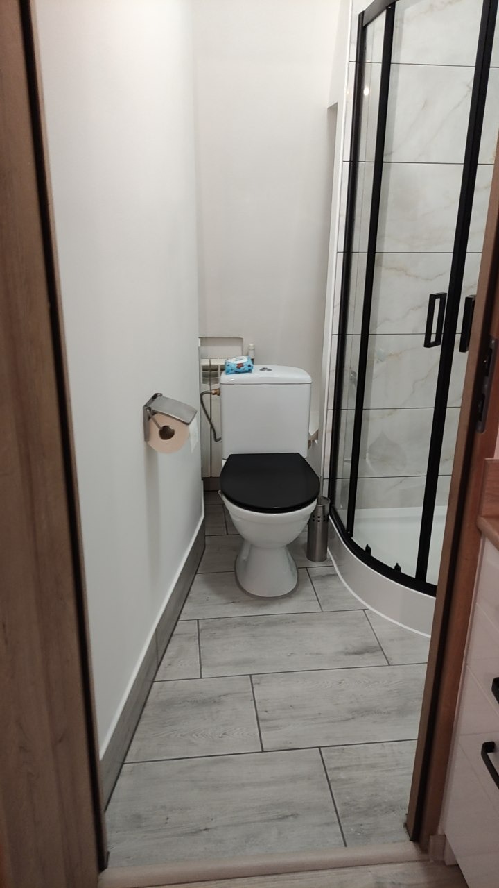 Nowoczesna łazienka z kabiną prysznicową typu walk-in, toaletą z czarną deską i szarą podłogą imitującą drewno. Widok zza drzwi.