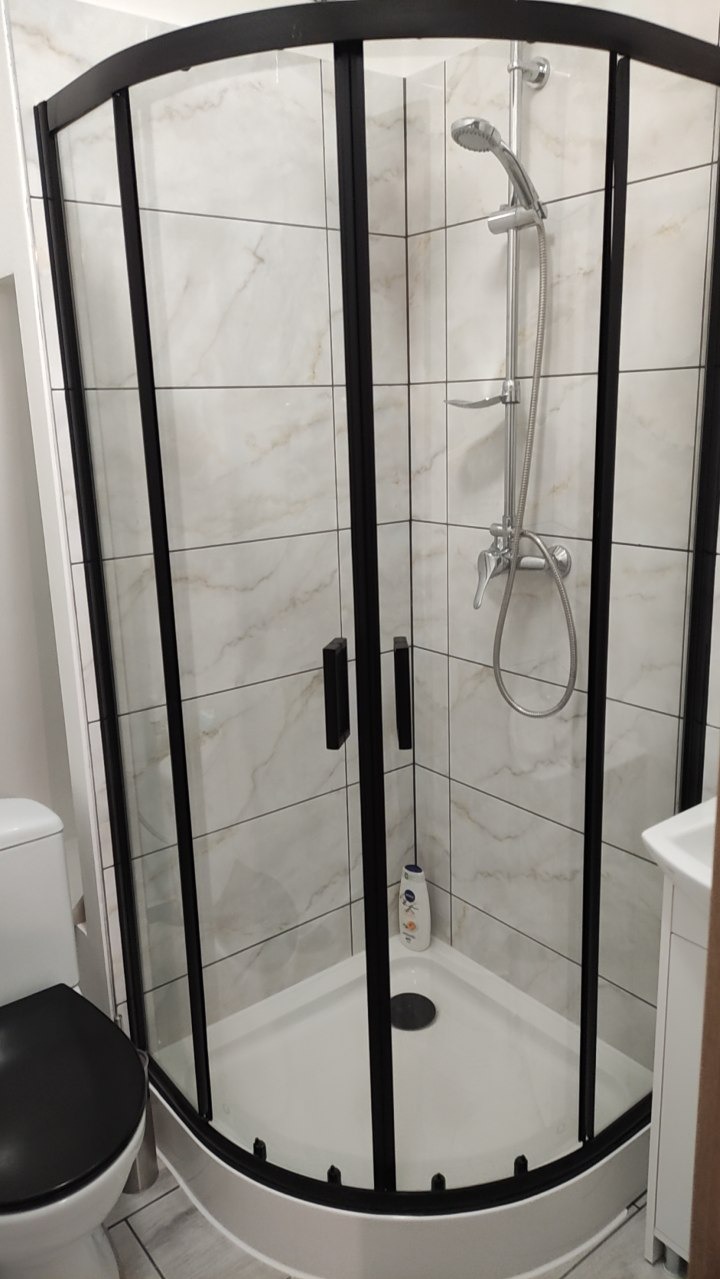 Narożna kabina prysznicowa z czarnymi profilami i marmurowymi płytkami w tle. Nowoczesne wykończenie łazienki po remoncie z widoczną armaturą.