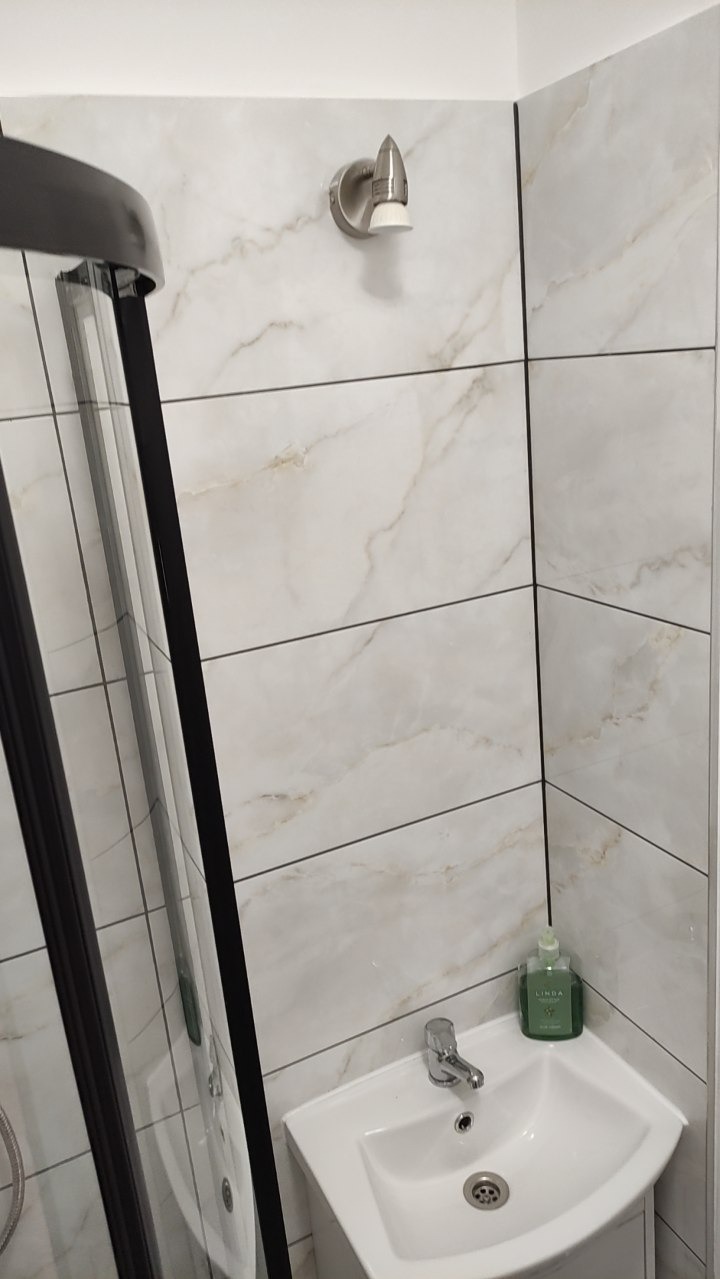 Wykończona łazienka z kabiną prysznicową, umywalką i baterią. Ściany wyłożone płytkami imitującymi marmur. Oświetlenie punktowe nad umywalką.