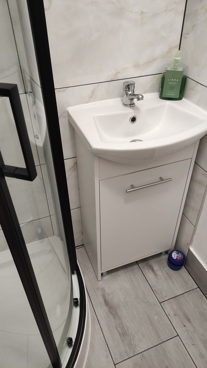 Biała umywalka z szafką i chromowaną baterią, obok kabina prysznicowa z czarnymi ramami, na tle marmurowych płytek. Minimalistyczna łazienka z szarymi płytkami podłogowymi.