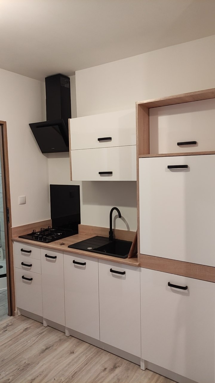 Nowoczesna, jasna kuchnia z białymi szafkami i drewnianym blatem. Czarny zlew i płyta gazowa dodają kontrastu. Widoczny minimalistyczny okap.