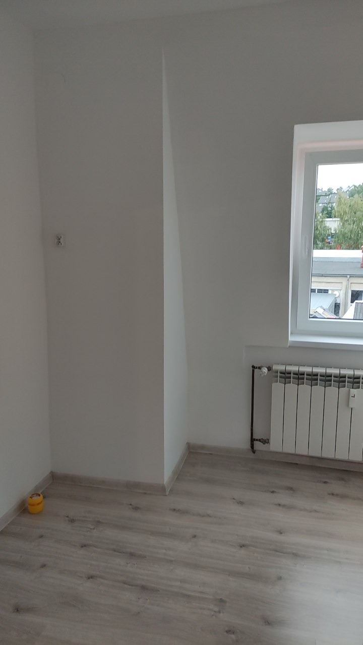 Jasne wnętrze po remoncie: białe ściany z grzejnikiem pod oknem, panele podłogowe. Widoczny mały, żółty pojemnik w rogu pokoju i elementy instalacji elektrycznej.
