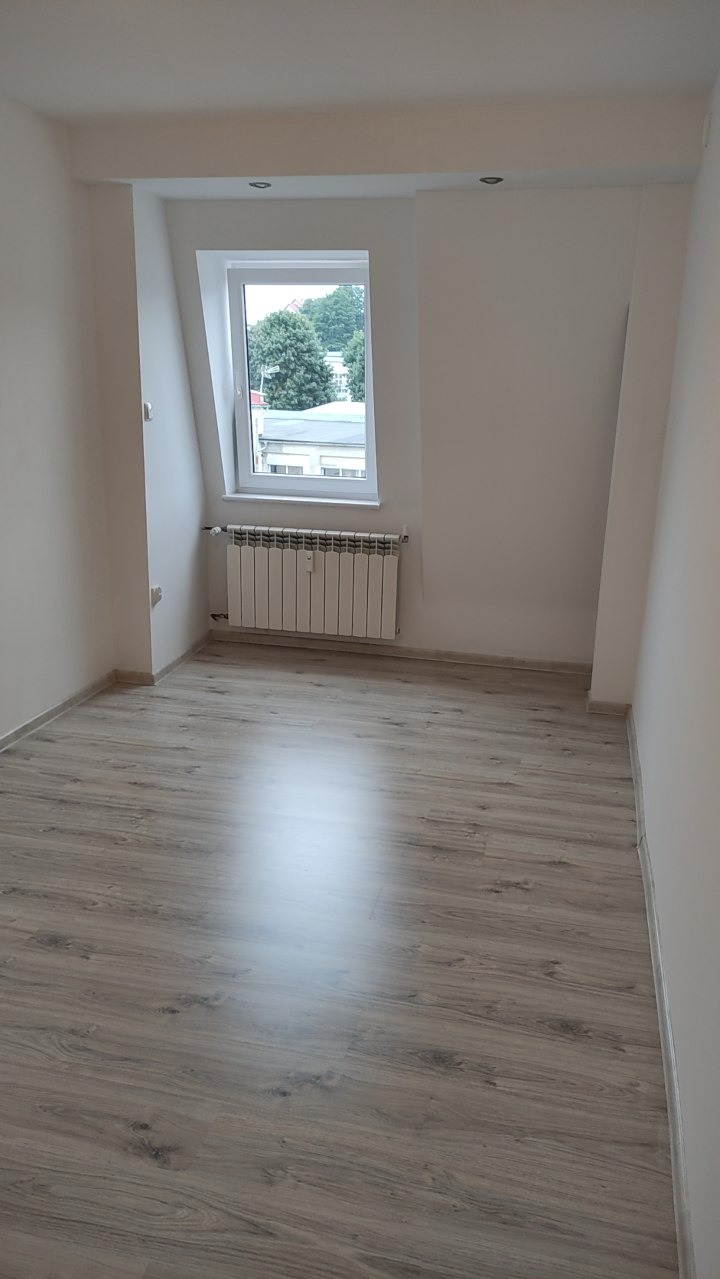 Jasne wnętrze po remoncie: panele podłogowe, białe ściany, okno dachowe z widokiem na zieleń. Nowy grzejnik pod oknem. Minimalistyczny, odświeżony pokój.
