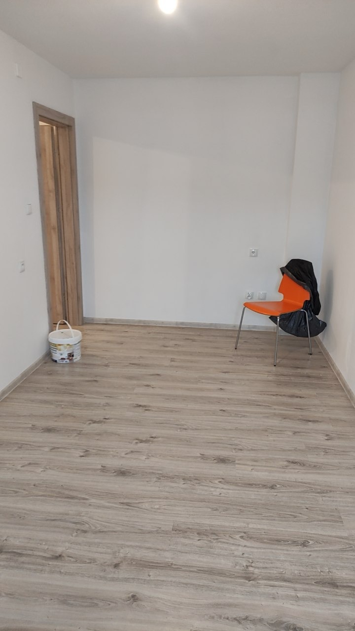Jasne panele podłogowe w pustym pokoju z pomarańczowym krzesłem i wiaderkiem farby przy drzwiach. Świeżo wykończone wnętrze, gotowe do umeblowania. Minimalistyczny wystrój.
