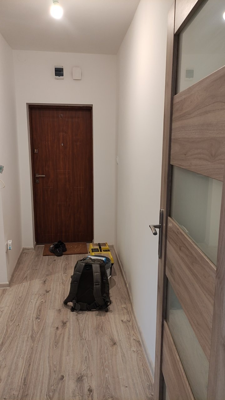 Korytarz po remoncie: drewnopodobna podłoga, białe ściany, brązowe drzwi wejściowe i wewnętrzne z przeszkleniami. Plecak i narzędzia na podłodze.