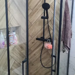 Kabina prysznicowa z czarnymi profilami na tle ściany z płytek imitujących drewno ułożonych w jodełkę, widoczny zestaw prysznicowy w kolorze czarnym, półka z kosmetykami i pralka.